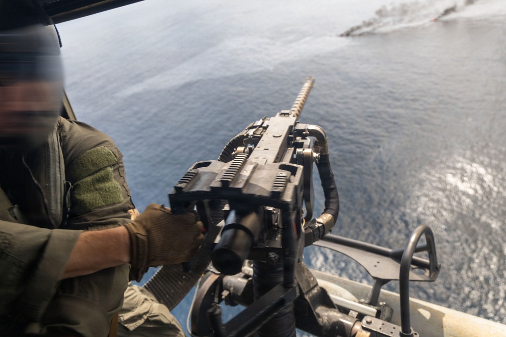 A military personnel is operating a door-mounted machine gun from what appears to be a helicopter flying over open ocean water. The gunner is wearing olive drab flight gear and tan tactical gloves while manning the weapon system. The perspective is from inside the aircraft looking outward toward the water below.