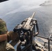 A military personnel is operating a door-mounted machine gun from what appears to be a helicopter flying over open ocean water. The gunner is wearing olive drab flight gear and tan tactical gloves while manning the weapon system. The perspective is from inside the aircraft looking outward toward the water below.