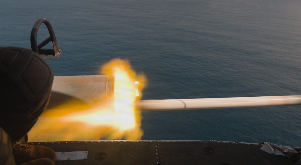 The image captures a weapon being fired from what appears to be a naval vessel, with a visible muzzle flash and projectile or barrel extending toward the right. The background shows open ocean water, suggesting this is a maritime military exercise or operation. A person in military attire is partially visible on the left side of the frame.