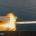 The image captures a weapon being fired from what appears to be a naval vessel, with a visible muzzle flash and projectile or barrel extending toward the right. The background shows open ocean water, suggesting this is a maritime military exercise or operation. A person in military attire is partially visible on the left side of the frame.