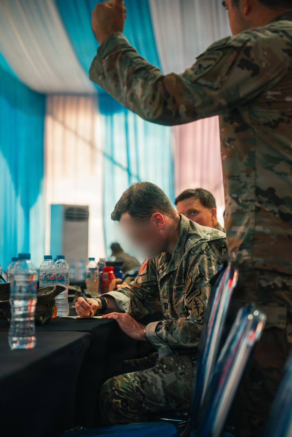 Military personnel in MultiCam/OCP camouflage uniforms are gathered in what appears to be a conference or briefing room. A seated soldier wearing U.S. Army patches is writing on documents at a table covered with a black cloth. The setting suggests a formal military planning session or coordination meeting.