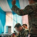 Military personnel in MultiCam/OCP camouflage uniforms are gathered in what appears to be a conference or briefing room. A seated soldier wearing U.S. Army patches is writing on documents at a table covered with a black cloth. The setting suggests a formal military planning session or coordination meeting.