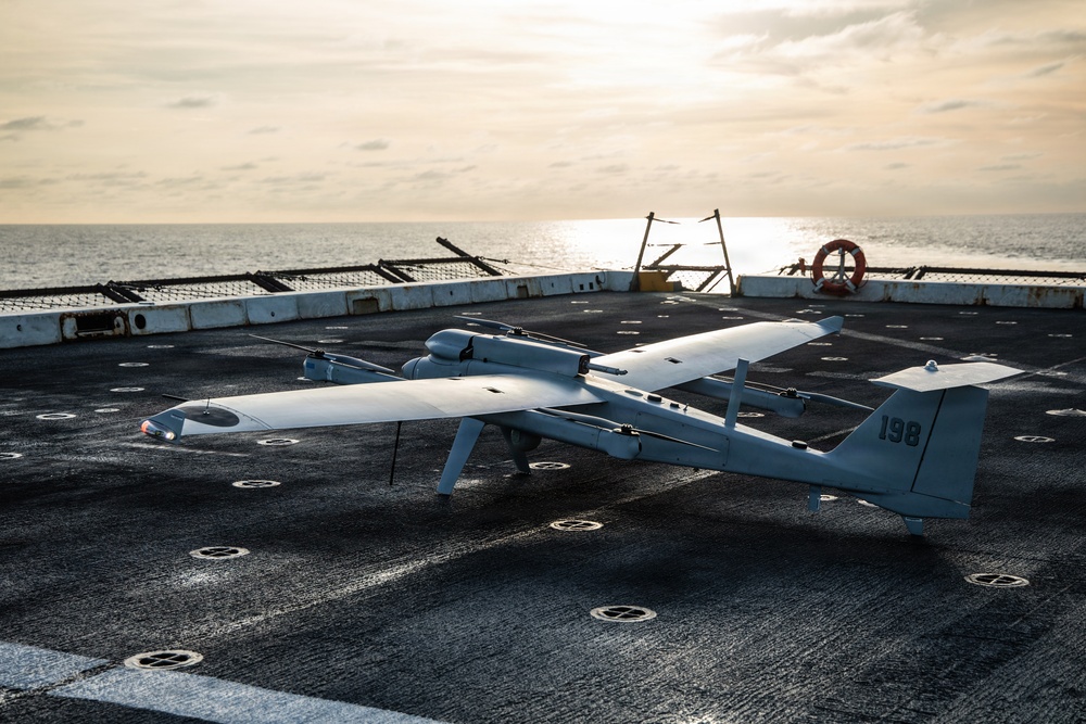 A military unmanned aerial vehicle (UAV) marked with the number '198' is parked on the flight deck of a naval vessel at sea. The drone appears to be a fixed-wing VTOL (Vertical Take-Off and Landing) type with multiple rotors mounted on the wings. The image is taken during golden hour with the ocean horizon visible in the background.
