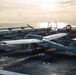 A military unmanned aerial vehicle (UAV) marked with the number '198' is parked on the flight deck of a naval vessel at sea. The drone appears to be a fixed-wing VTOL (Vertical Take-Off and Landing) type with multiple rotors mounted on the wings. The image is taken during golden hour with the ocean horizon visible in the background.