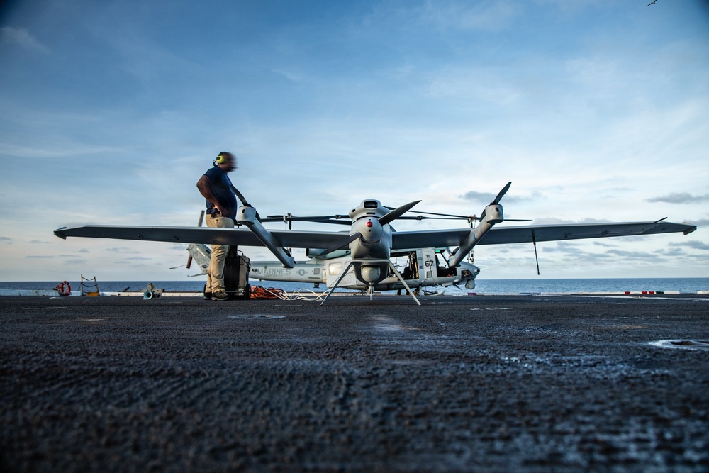 A military technician is performing pre-flight or maintenance checks on a large unmanned aerial vehicle (UAV) on the flight deck of a naval vessel at sea. The aircraft bears 'MARINES' markings and the number '67', consistent with a U.S. Marine Corps drone such as the MQ-9 Reaper or similar MALE UAV. The scene is captured at low angle during overcast but bright conditions with open ocean visible in the background.