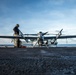 A military technician is performing pre-flight or maintenance checks on a large unmanned aerial vehicle (UAV) on the flight deck of a naval vessel at sea. The aircraft bears 'MARINES' markings and the number '67', consistent with a U.S. Marine Corps drone such as the MQ-9 Reaper or similar MALE UAV. The scene is captured at low angle during overcast but bright conditions with open ocean visible in the background.