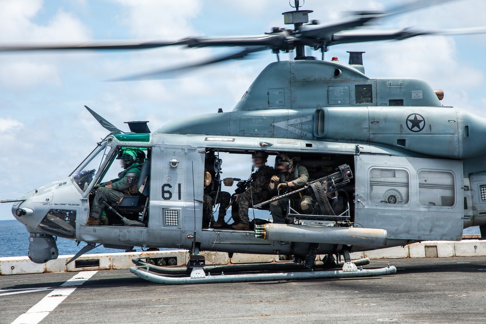 A U.S. military helicopter, numbered '61', is on the deck of a naval vessel with rotors spinning. Several military personnel are visible inside the open cabin, with at least one crew member manning a door-mounted machine gun. The aircraft bears U.S. military markings including a star insignia and appears to be a UH-1Y Venom or similar utility helicopter.