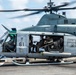 A U.S. military helicopter, numbered '61', is on the deck of a naval vessel with rotors spinning. Several military personnel are visible inside the open cabin, with at least one crew member manning a door-mounted machine gun. The aircraft bears U.S. military markings including a star insignia and appears to be a UH-1Y Venom or similar utility helicopter.