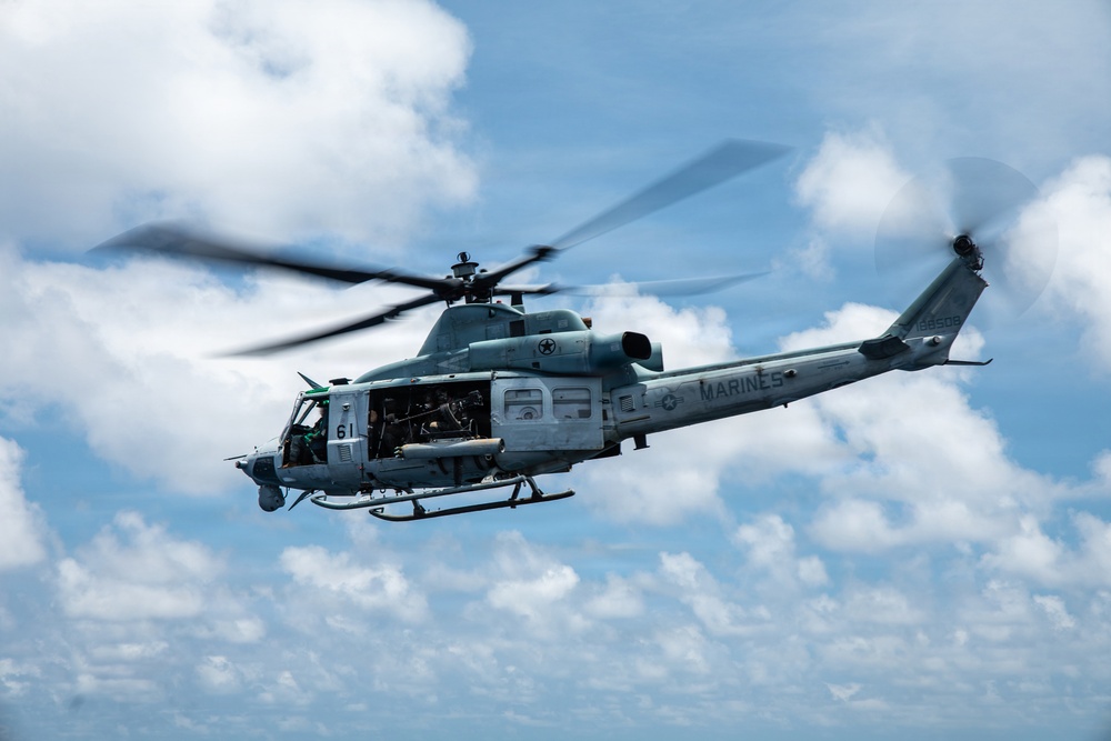 A U.S. Marines military helicopter is captured in flight against a partly cloudy blue sky. The aircraft is a light gray utility/attack helicopter with the word 'MARINES' clearly stenciled on the fuselage, bearing the number '61' on its nose section. The rotor blades show motion blur, indicating active flight.