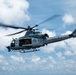 A U.S. Marines military helicopter is captured in flight against a partly cloudy blue sky. The aircraft is a light gray utility/attack helicopter with the word 'MARINES' clearly stenciled on the fuselage, bearing the number '61' on its nose section. The rotor blades show motion blur, indicating active flight.