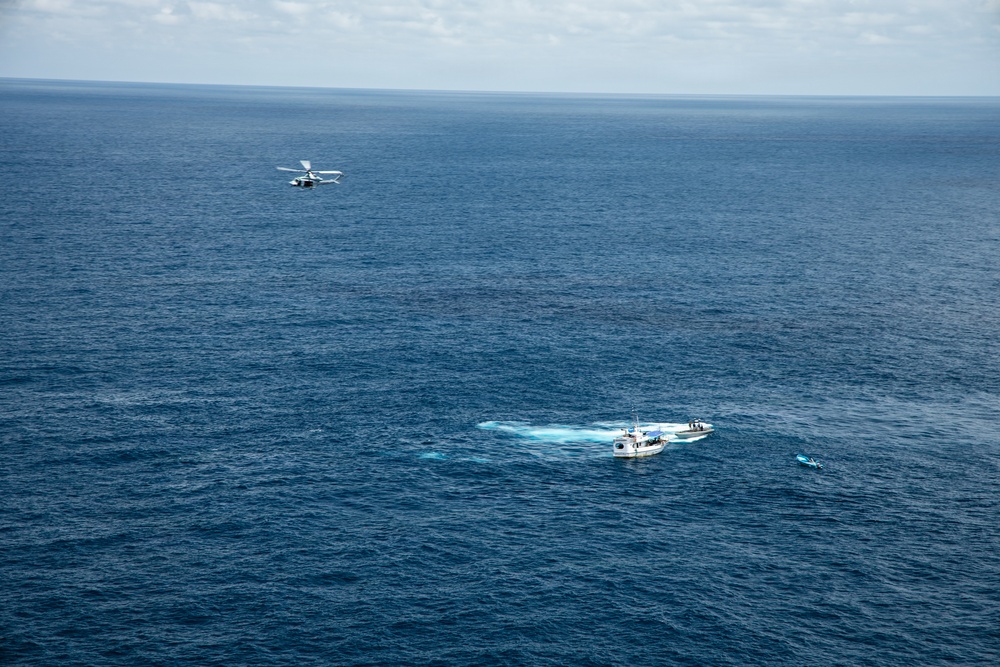 An aerial photograph taken over open ocean showing a white vessel and a small aircraft in proximity to each other. The boat appears to be stationary or moving slowly, with a visible wake/disturbance in the water nearby. A smaller object, possibly a kayak or small watercraft, is visible to the right of the main vessel.