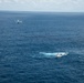 An aerial photograph taken over open ocean showing a white vessel and a small aircraft in proximity to each other. The boat appears to be stationary or moving slowly, with a visible wake/disturbance in the water nearby. A smaller object, possibly a kayak or small watercraft, is visible to the right of the main vessel.
