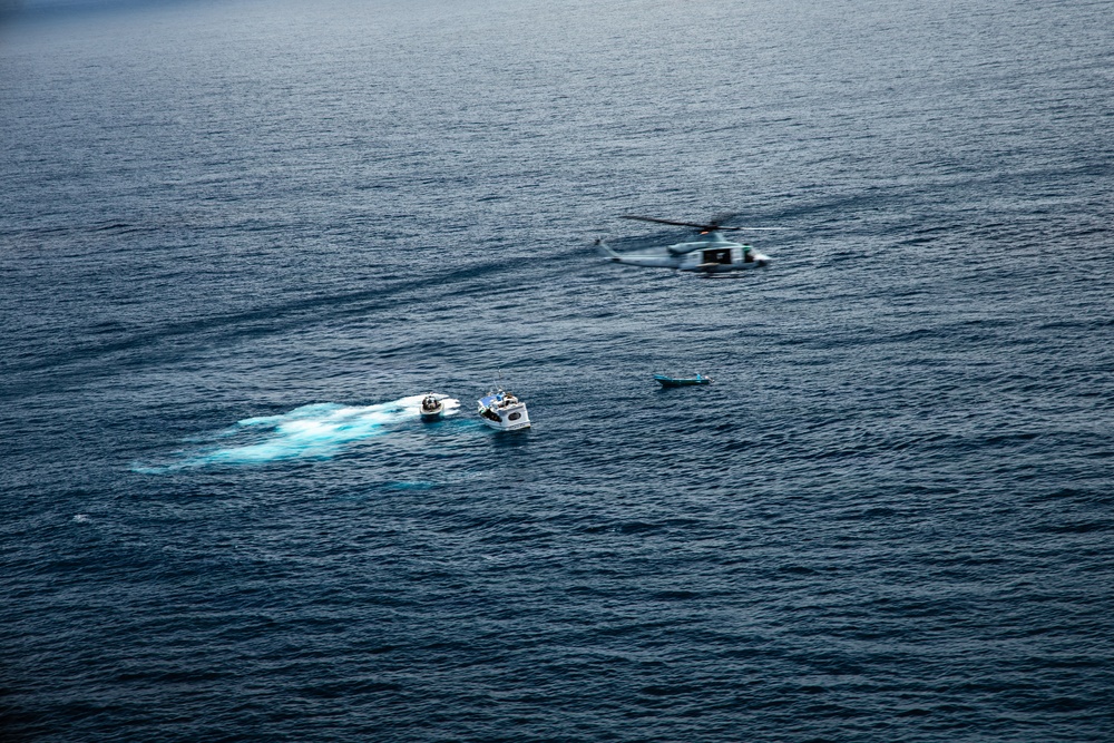 An aerial photograph taken over open ocean showing a maritime operation involving two motorboats and a helicopter. One white motorboat appears to be intercepting or alongside a smaller dark vessel, while a military or coast guard helicopter hovers nearby. A third small vessel or object is visible slightly further away on the water.