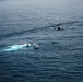 An aerial photograph taken over open ocean showing a maritime operation involving two motorboats and a helicopter. One white motorboat appears to be intercepting or alongside a smaller dark vessel, while a military or coast guard helicopter hovers nearby. A third small vessel or object is visible slightly further away on the water.