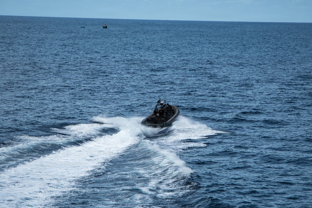 A rigid inflatable boat (RIB) is traveling at high speed across open ocean, generating a prominent white wake. The vessel appears to be a military or law enforcement type craft with personnel and equipment visible on board. A second, smaller vessel is visible in the far background near the horizon.