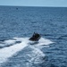 A rigid inflatable boat (RIB) is traveling at high speed across open ocean, generating a prominent white wake. The vessel appears to be a military or law enforcement type craft with personnel and equipment visible on board. A second, smaller vessel is visible in the far background near the horizon.