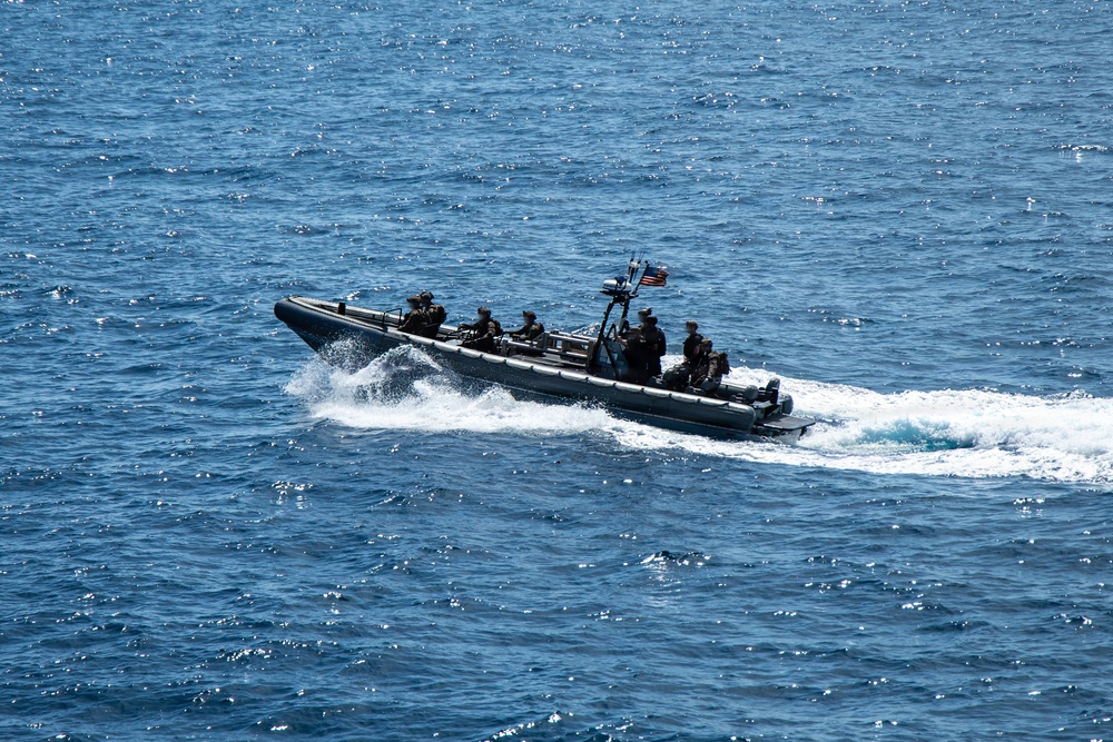 A military rigid inflatable boat (RIB) is underway at speed on open blue ocean water. The vessel carries multiple personnel in dark tactical gear and displays a visible American flag at the stern. The boat is creating a prominent white wake, indicating high-speed travel.