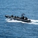 A military rigid inflatable boat (RIB) is underway at speed on open blue ocean water. The vessel carries multiple personnel in dark tactical gear and displays a visible American flag at the stern. The boat is creating a prominent white wake, indicating high-speed travel.