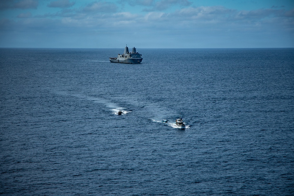 A large naval warship, consistent with an amphibious transport dock vessel, is positioned on the horizon while three smaller military patrol or rigid-hull inflatable boats (RHIBs) operate in the foreground, moving toward or away from the larger vessel. The scene appears to depict a maritime military exercise or small boat operations in open ocean. The image was taken from an elevated vantage point, likely from another vessel or aircraft.