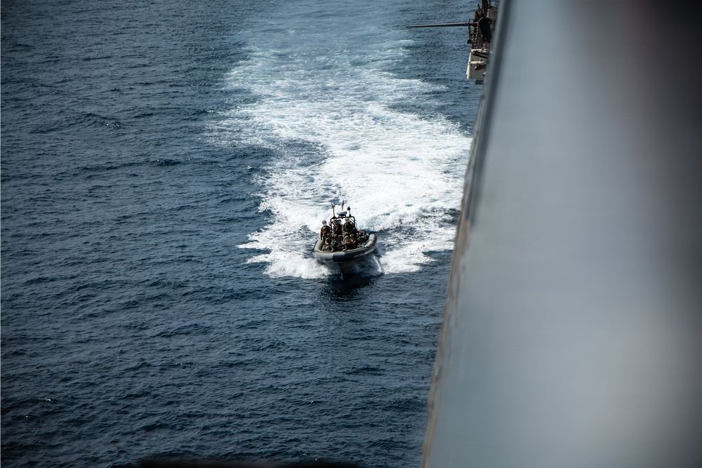 An aerial or elevated view of a rigid inflatable boat (RIB) traveling at speed alongside a large naval vessel in open ocean. The small boat carries multiple personnel and is leaving a prominent white wake behind it. The photograph appears to be taken from the deck or superstructure of the larger ship.