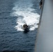 An aerial or elevated view of a rigid inflatable boat (RIB) traveling at speed alongside a large naval vessel in open ocean. The small boat carries multiple personnel and is leaving a prominent white wake behind it. The photograph appears to be taken from the deck or superstructure of the larger ship.