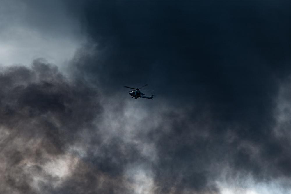 A helicopter is captured in flight against a dramatically dark and turbulent sky filled with dense, billowing clouds that appear to be smoke or storm clouds. The aircraft is silhouetted against the dark background, positioned near the center of the frame. The overall atmosphere is ominous and tense, suggesting either a storm environment or proximity to a large fire or explosion.