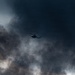 A helicopter is captured in flight against a dramatically dark and turbulent sky filled with dense, billowing clouds that appear to be smoke or storm clouds. The aircraft is silhouetted against the dark background, positioned near the center of the frame. The overall atmosphere is ominous and tense, suggesting either a storm environment or proximity to a large fire or explosion.