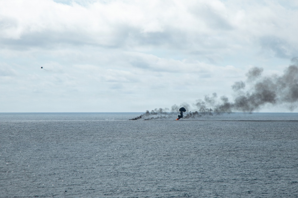 The image shows a large fire burning on the open ocean surface, producing thick black smoke that billows upward and spreads horizontally across the horizon. A small airborne object, likely a helicopter or aircraft, is visible in the upper left portion of the sky. The scene suggests a maritime incident, controlled burn, or military exercise taking place at sea.