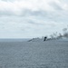 The image shows a large fire burning on the open ocean surface, producing thick black smoke that billows upward and spreads horizontally across the horizon. A small airborne object, likely a helicopter or aircraft, is visible in the upper left portion of the sky. The scene suggests a maritime incident, controlled burn, or military exercise taking place at sea.