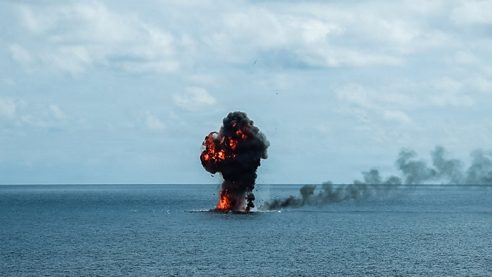 The image shows a large explosion occurring on the surface of an open body of water, producing a significant fireball with orange and red flames accompanied by dense black smoke rising into the overcast sky. A secondary smoke trail is visible drifting to the right along the horizon. The scene appears to depict a maritime detonation or vessel destruction event.