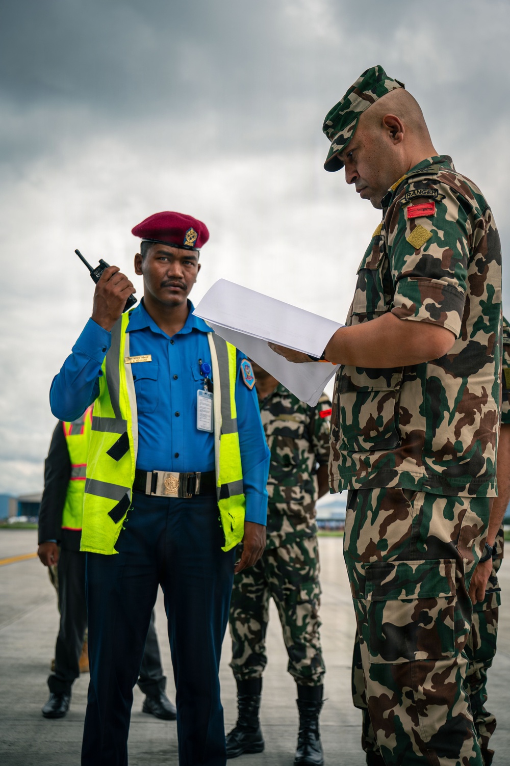 The image shows a military officer in camouflage uniform reviewing documents while interacting with a security/police officer wearing a blue shirt, high-visibility vest, and maroon beret on what appears to be an airport tarmac. The military officer has a 'RANGER' tab visible on his shoulder. Additional personnel in camouflage uniforms are visible in the background.