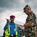 The image shows a military officer in camouflage uniform reviewing documents while interacting with a security/police officer wearing a blue shirt, high-visibility vest, and maroon beret on what appears to be an airport tarmac. The military officer has a 'RANGER' tab visible on his shoulder. Additional personnel in camouflage uniforms are visible in the background.