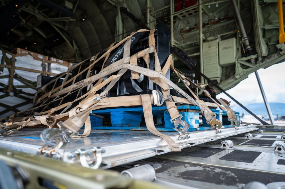 The image shows the interior cargo bay of a military transport aircraft, likely a C-130 Hercules, with a secured cargo pallet loaded on the roller-equipped floor. The cargo is strapped down with multiple tan/beige military-grade tie-down straps and cargo nets attached to anchor points along the aircraft floor. The load appears to be positioned near the rear ramp area of the aircraft, ready for transport or airdrop operations.