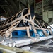 The image shows the interior cargo bay of a military transport aircraft, likely a C-130 Hercules, with a secured cargo pallet loaded on the roller-equipped floor. The cargo is strapped down with multiple tan/beige military-grade tie-down straps and cargo nets attached to anchor points along the aircraft floor. The load appears to be positioned near the rear ramp area of the aircraft, ready for transport or airdrop operations.