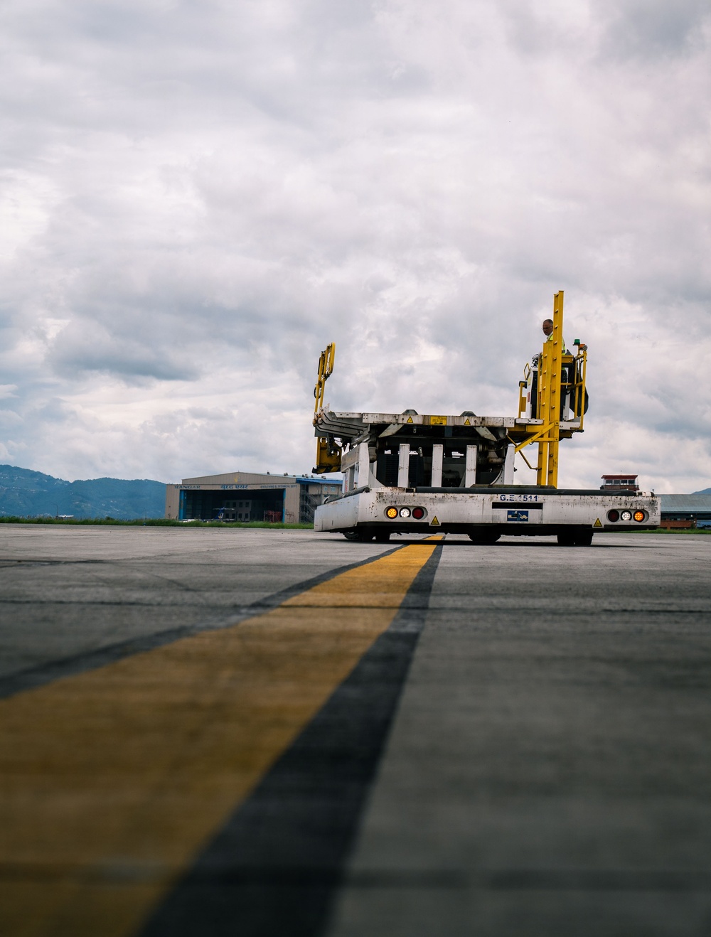 A ground support vehicle, specifically an aircraft loader or cargo lift truck, is positioned on an airport apron/taxiway. The vehicle bears the marking 'G.E. 511' on its rear and features yellow structural framework with a raised platform. Airport hangars and mountainous terrain are visible in the background under an overcast sky.