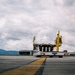 A ground support vehicle, specifically an aircraft loader or cargo lift truck, is positioned on an airport apron/taxiway. The vehicle bears the marking 'G.E. 511' on its rear and features yellow structural framework with a raised platform. Airport hangars and mountainous terrain are visible in the background under an overcast sky.