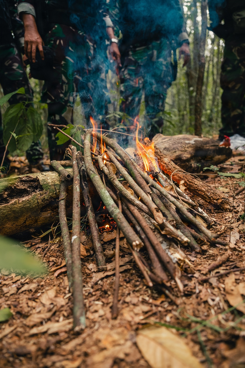 A small campfire built from bundled sticks and branches is actively burning on a forest floor covered with dry leaves and debris. Two individuals wearing military-style camouflage uniforms are standing behind the fire, visible from the waist down. The setting appears to be a dense tropical or subtropical forest, suggesting a field training or survival exercise.