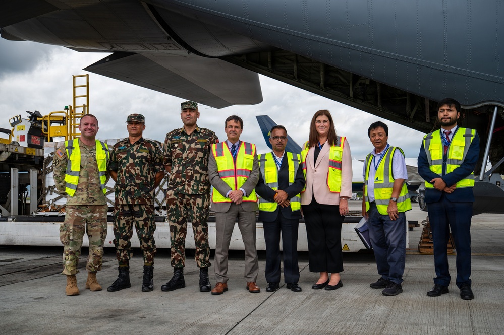 A group of eight individuals pose for a formal photograph on an airport tarmac beneath the wing of a large military or cargo aircraft. The group includes military personnel in camouflage uniforms, civilians in business attire, and airport/logistics staff wearing high-visibility safety vests. The setting suggests an official handover, humanitarian aid delivery, or bilateral military cooperation event.