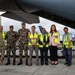 A group of eight individuals pose for a formal photograph on an airport tarmac beneath the wing of a large military or cargo aircraft. The group includes military personnel in camouflage uniforms, civilians in business attire, and airport/logistics staff wearing high-visibility safety vests. The setting suggests an official handover, humanitarian aid delivery, or bilateral military cooperation event.