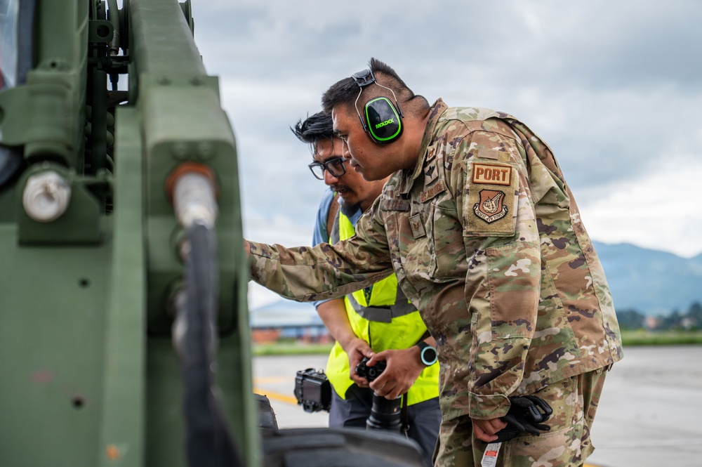 A U.S. Air Force service member wearing OCP (Operational Camouflage Pattern) uniform with a 'PORT' tab and green MOLDEX hearing protection is inspecting or working on military equipment at an airfield. A second individual wearing a high-visibility yellow vest and glasses stands nearby, holding a camera. The setting appears to be an outdoor tarmac or flight line with mountains and overcast skies visible in the background.