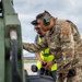 A U.S. Air Force service member wearing OCP (Operational Camouflage Pattern) uniform with a 'PORT' tab and green MOLDEX hearing protection is inspecting or working on military equipment at an airfield. A second individual wearing a high-visibility yellow vest and glasses stands nearby, holding a camera. The setting appears to be an outdoor tarmac or flight line with mountains and overcast skies visible in the background.