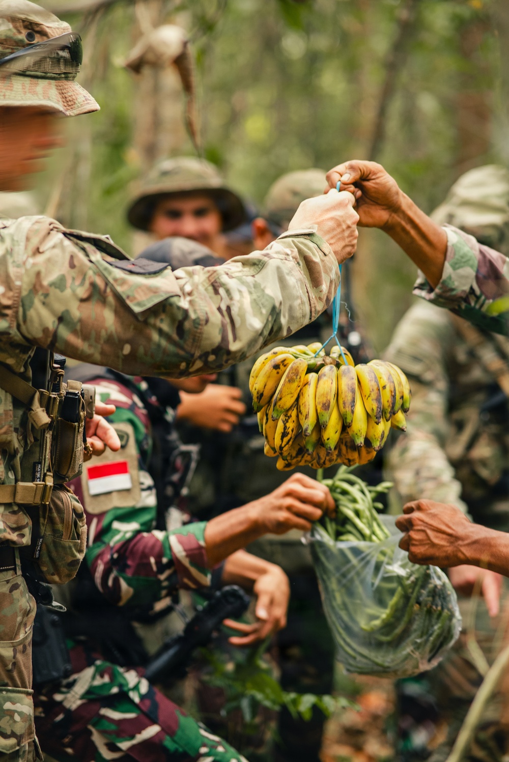 Military personnel in a jungle or forested environment are exchanging or receiving fresh produce, including a bunch of bananas and green vegetables. At least two distinct camouflage uniforms are visible, one bearing an Indonesian flag patch (red and white), suggesting a joint or multinational military exercise. The interaction appears cooperative and informal in nature.