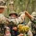 Military personnel in a jungle or forested environment are exchanging or receiving fresh produce, including a bunch of bananas and green vegetables. At least two distinct camouflage uniforms are visible, one bearing an Indonesian flag patch (red and white), suggesting a joint or multinational military exercise. The interaction appears cooperative and informal in nature.