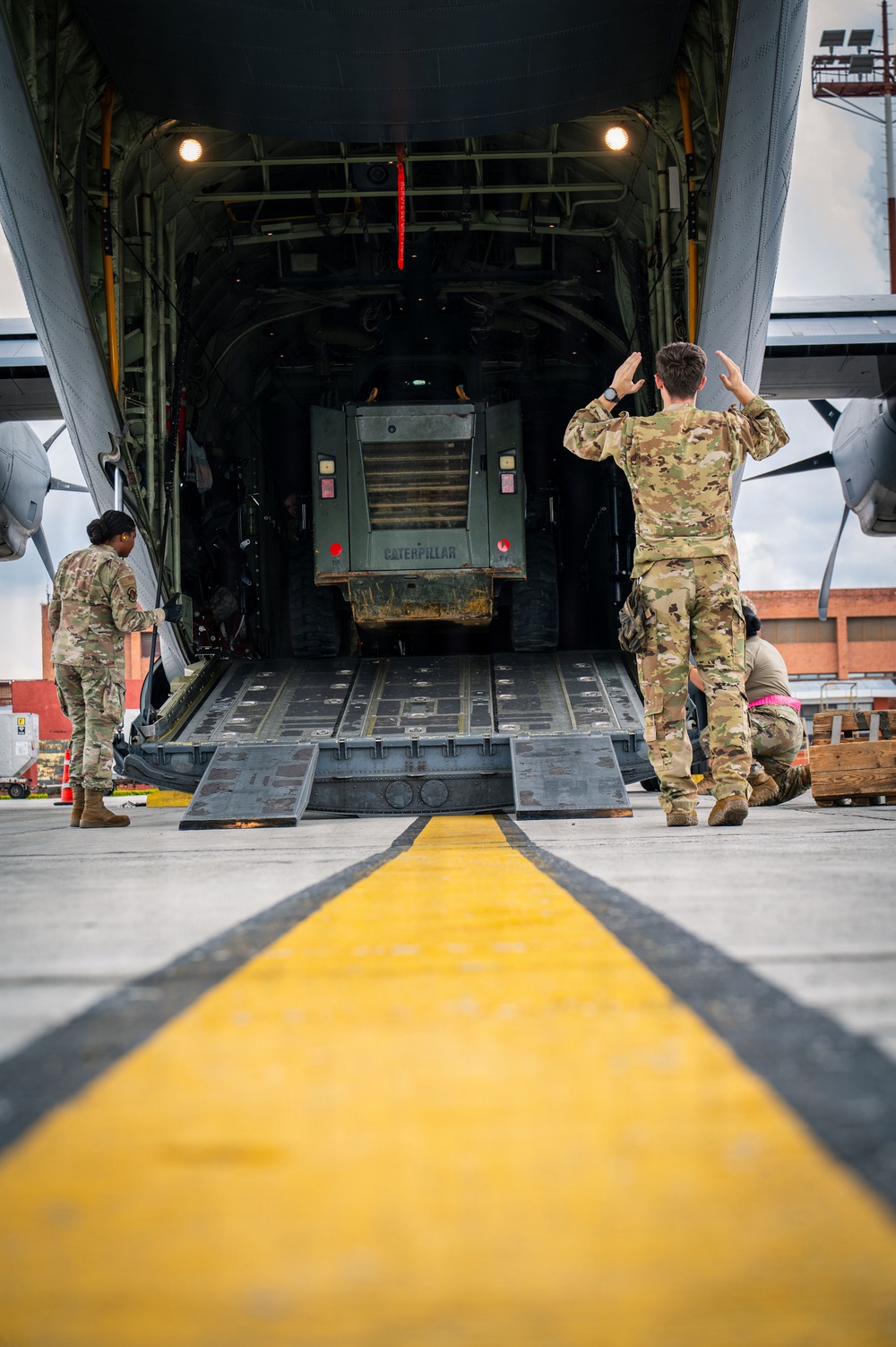 Military personnel in OCP (Operational Camouflage Pattern) uniforms are loading a Caterpillar vehicle into the rear cargo ramp of a large military transport aircraft, likely a C-17 Globemaster or C-130. One service member is using hand signals to guide the vehicle up the ramp while others assist on the sides. The scene takes place on a tarmac marked with yellow safety lines.