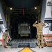 Military personnel in OCP (Operational Camouflage Pattern) uniforms are loading a Caterpillar vehicle into the rear cargo ramp of a large military transport aircraft, likely a C-17 Globemaster or C-130. One service member is using hand signals to guide the vehicle up the ramp while others assist on the sides. The scene takes place on a tarmac marked with yellow safety lines.