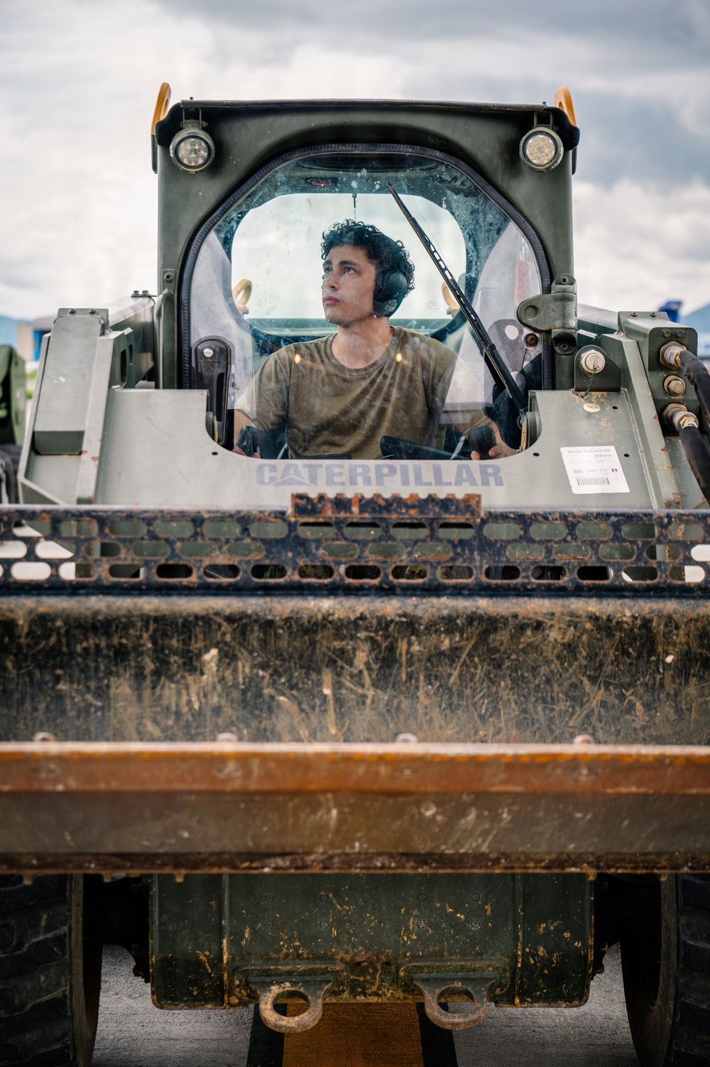 A young male operator is seated inside the enclosed cab of a Caterpillar skid steer loader or compact track loader, painted in olive/military green. He is wearing hearing protection (ear muffs) and a olive-colored t-shirt, suggesting a military or heavy construction context. The machine's bucket attachment is raised and visible in the foreground, showing significant dirt and wear.