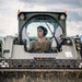 A young male operator is seated inside the enclosed cab of a Caterpillar skid steer loader or compact track loader, painted in olive/military green. He is wearing hearing protection (ear muffs) and a olive-colored t-shirt, suggesting a military or heavy construction context. The machine's bucket attachment is raised and visible in the foreground, showing significant dirt and wear.