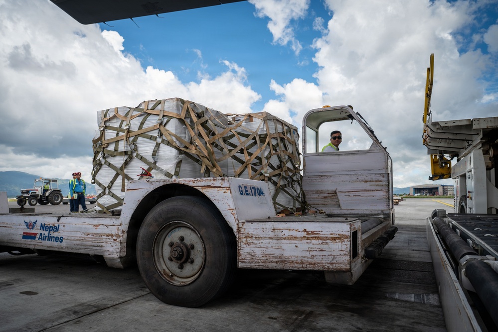 A Nepal Airlines ground support vehicle (unit G.E.1704) is transporting a large netted cargo pallet on an airport tarmac. Ground crew members in high-visibility vests are visible overseeing the cargo handling operation. The scene appears to be at Tribhuvan International Airport in Kathmandu, Nepal.