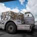 A Nepal Airlines ground support vehicle (unit G.E.1704) is transporting a large netted cargo pallet on an airport tarmac. Ground crew members in high-visibility vests are visible overseeing the cargo handling operation. The scene appears to be at Tribhuvan International Airport in Kathmandu, Nepal.