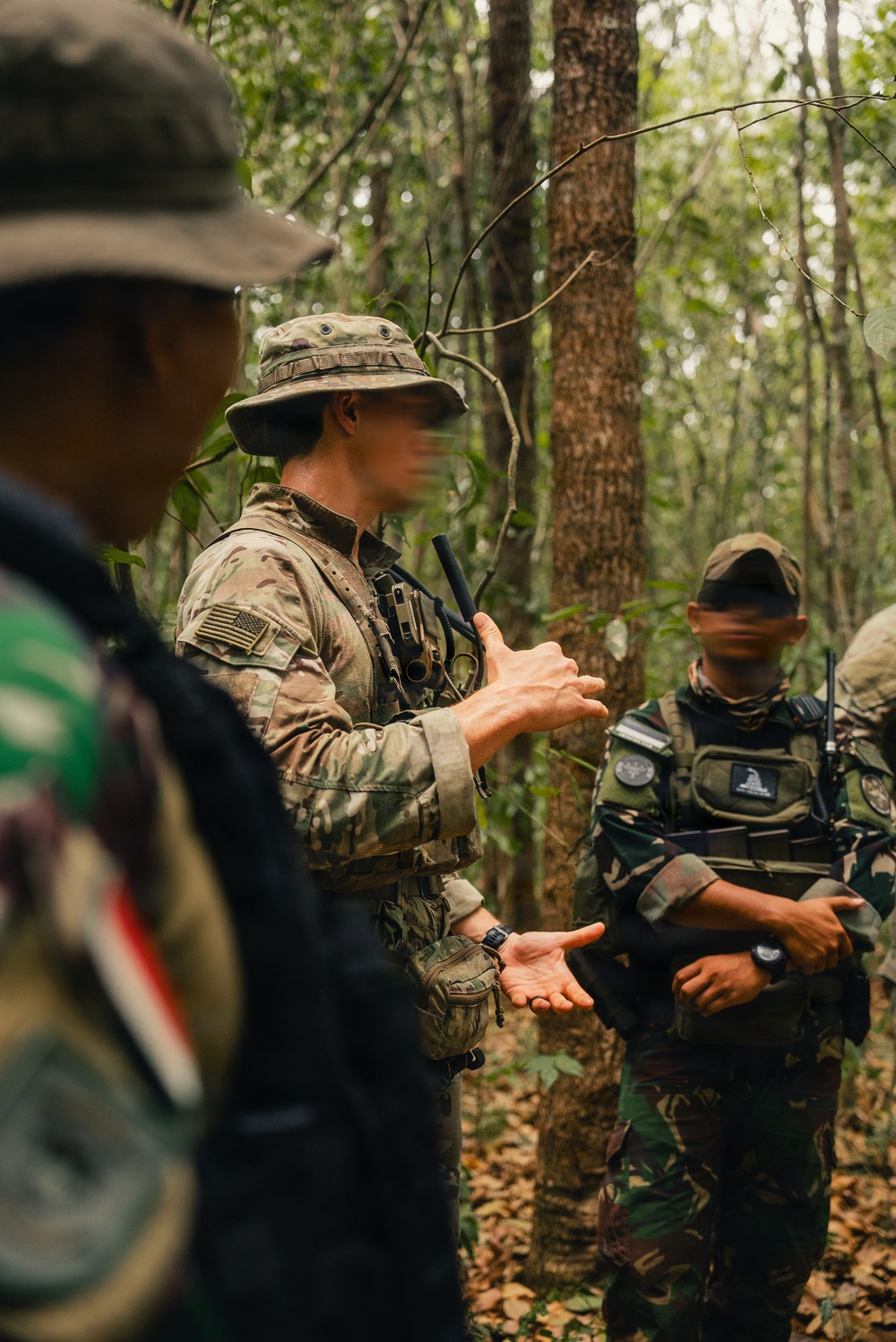 The image shows military personnel in a forested environment, appearing to conduct a briefing or instruction session. A central figure wearing MultiCam/OCP camouflage with a US flag patch and boonie hat is gesturing with their hands, apparently explaining something to others. The setting and uniforms suggest a joint training exercise involving at least two different military forces.